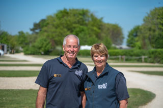 A man and woman in navy blue poloshirts smile at the camera. Behind them in the sunshine are premium pitches at Cotswold Farm Park Holidays